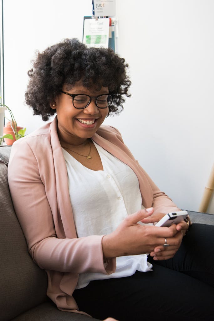 Joyful woman relaxing on a couch with a smartphone, embracing casual fashion and a bright smile.